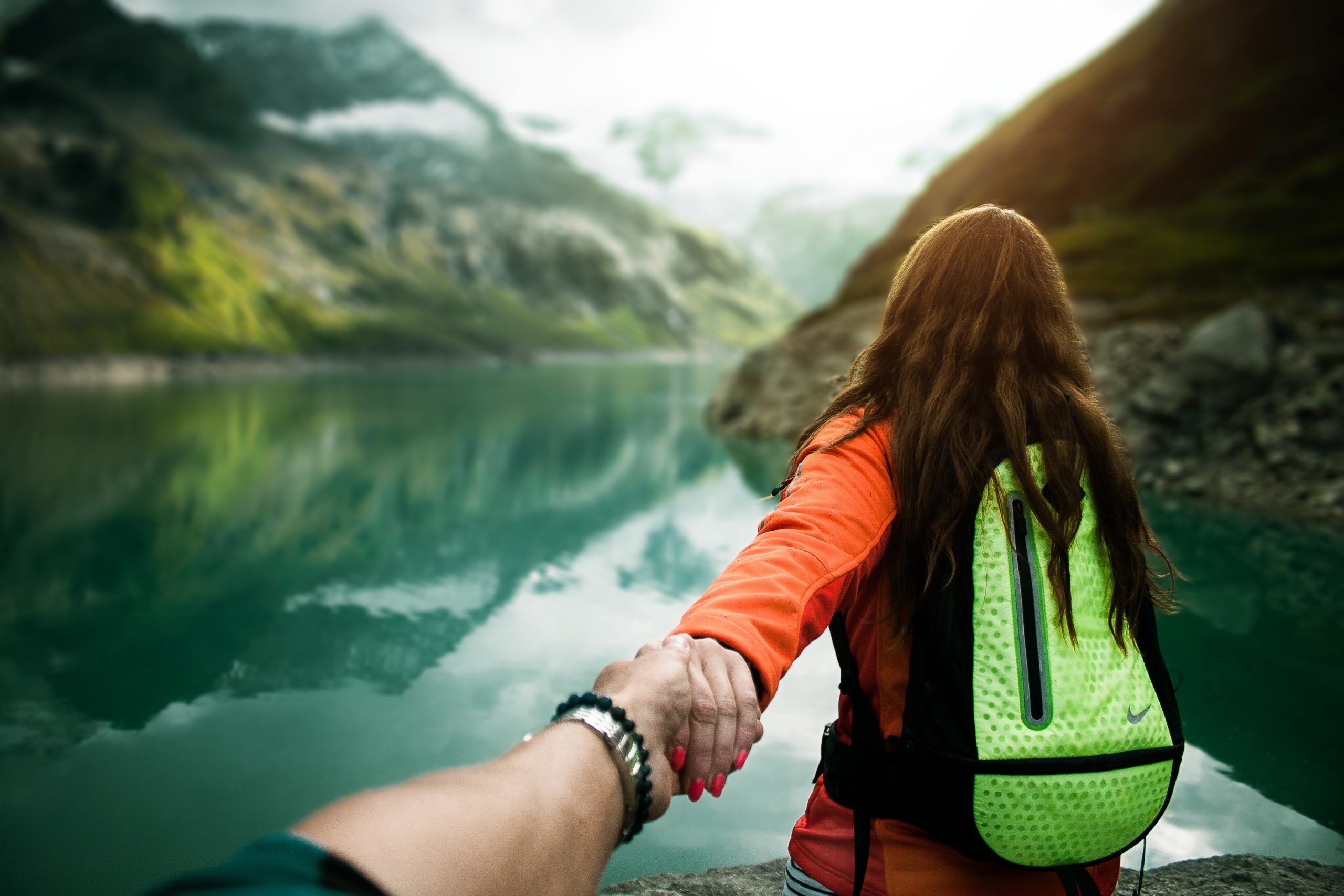 A person leads another by the hand toward a misty mountain lake