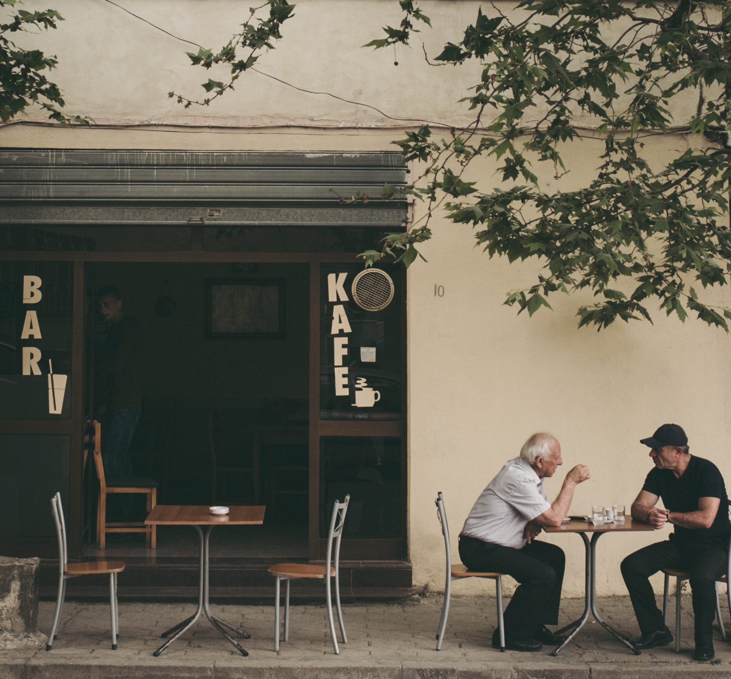 Two older men sit at a small café table outdoors, absorbed in conversation under a tree