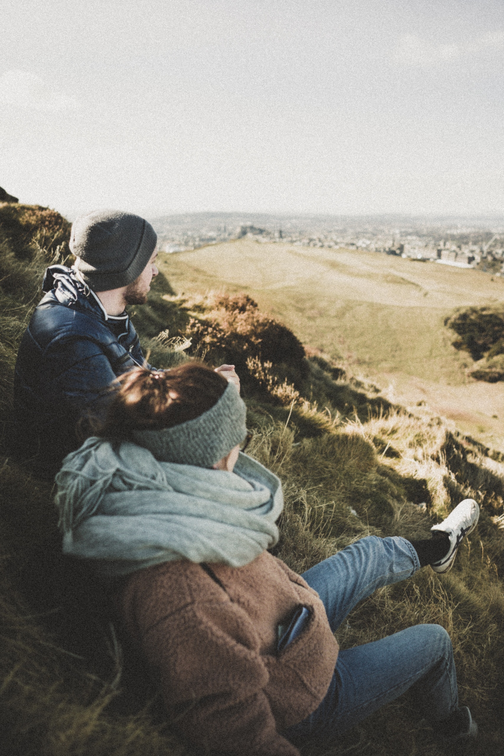 Two people rest on a grassy hillside overlooking a valley and distant city